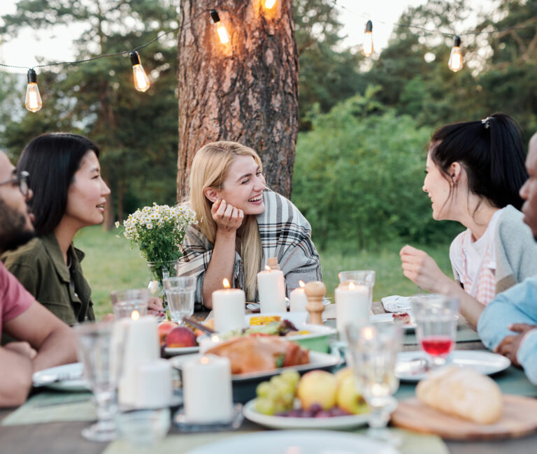 Young joyful couples sitting by served festive table under pine tree, talking, laughing and enjoying tasty food at outdoor dinner or party